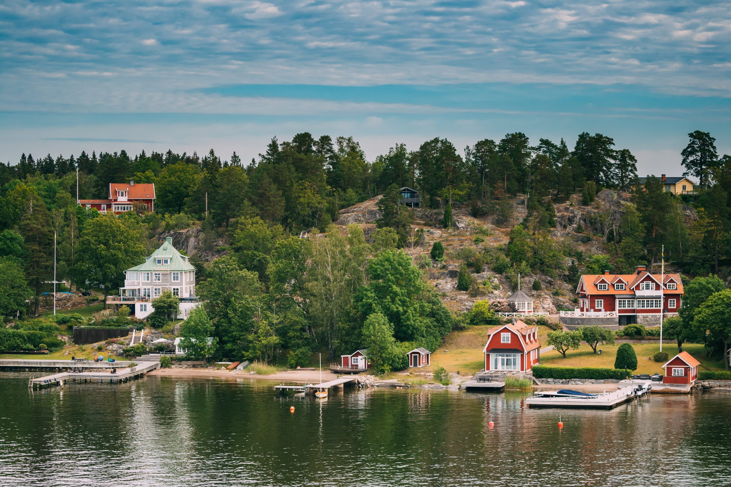 Sweden. Many Beautiful Red Swedish Wooden Log Cabins Houses On Rocky Island Coast In Summer Sunny Evening. Lake Or River Landscape.